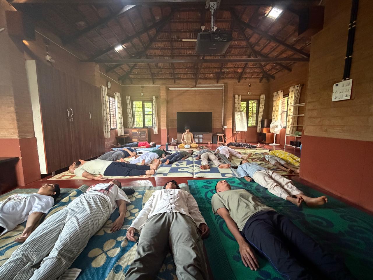 A large group resting in shavasana on mats in a rustic hall, Likhitha leading from the front