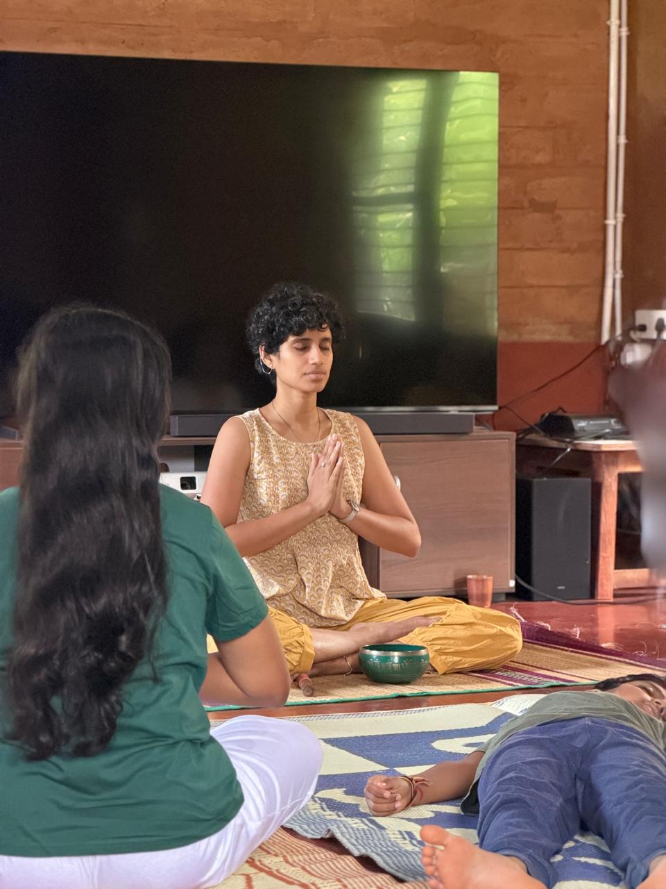 Likhitha seated in namaste with a singing bowl, leading a small group in meditation