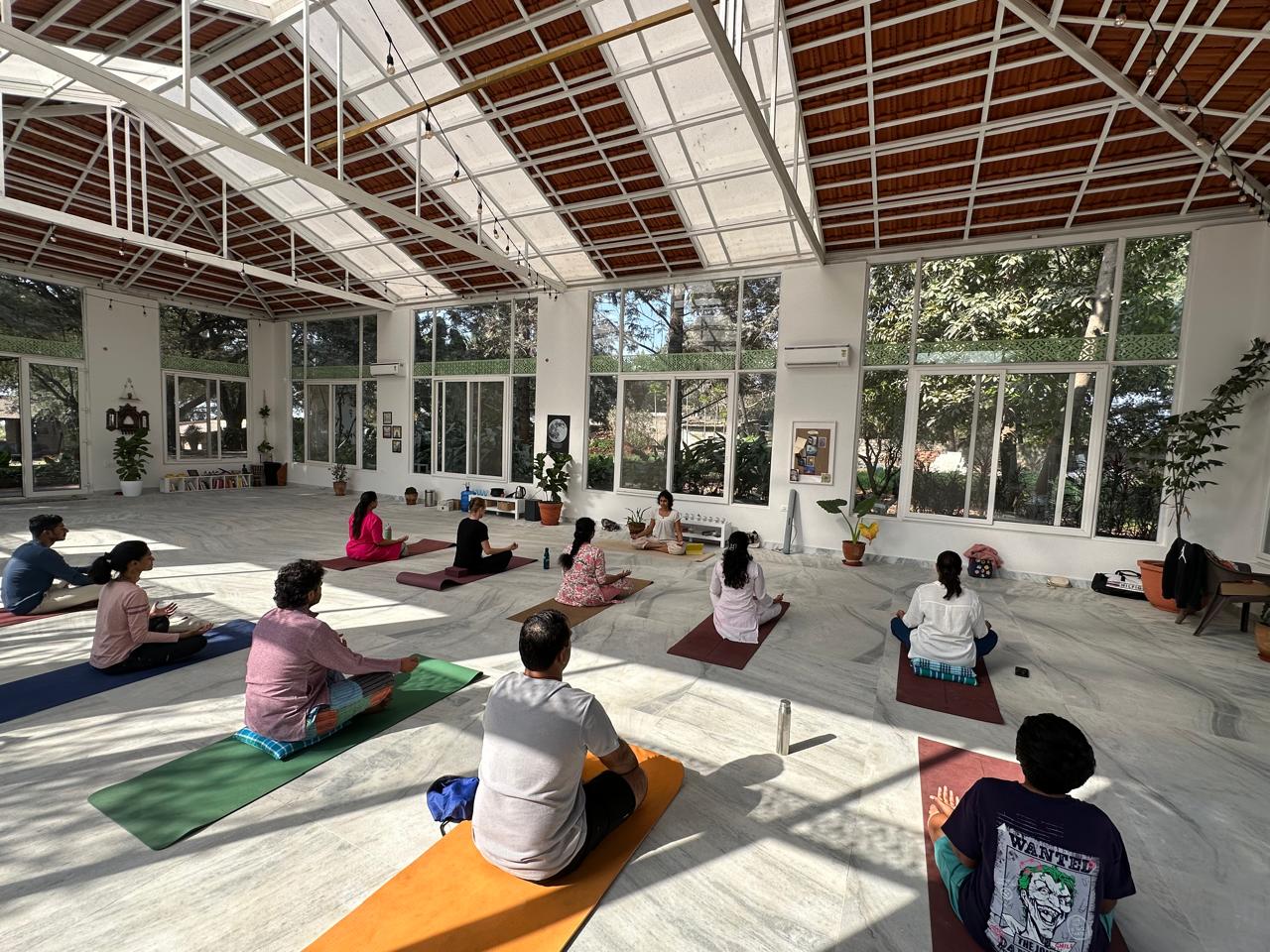 Group seated in meditation in a bright glass-roofed studio, Likhitha leading from the front