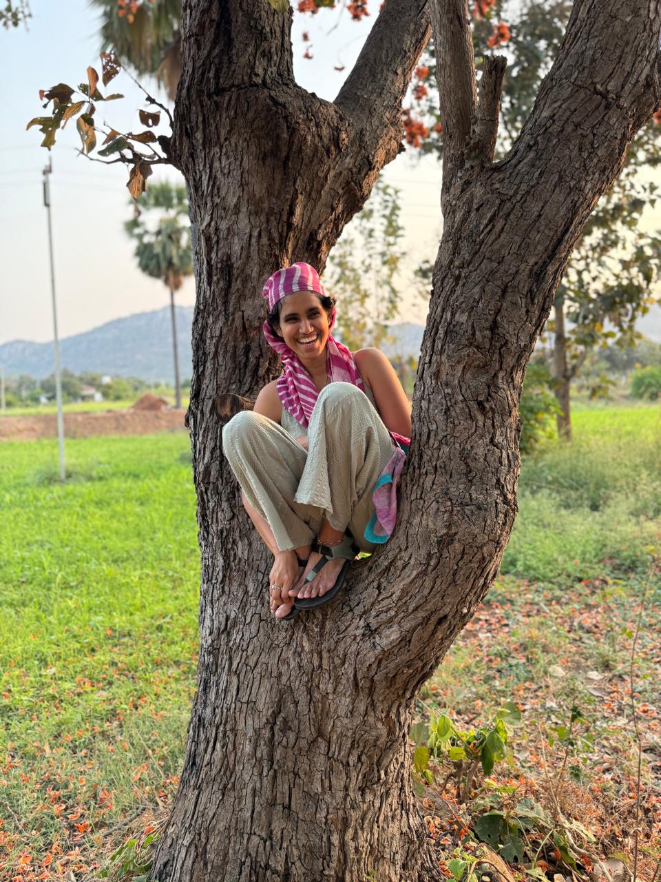 Likhitha sitting playfully in the fork of a tree, smiling, a rural field behind her