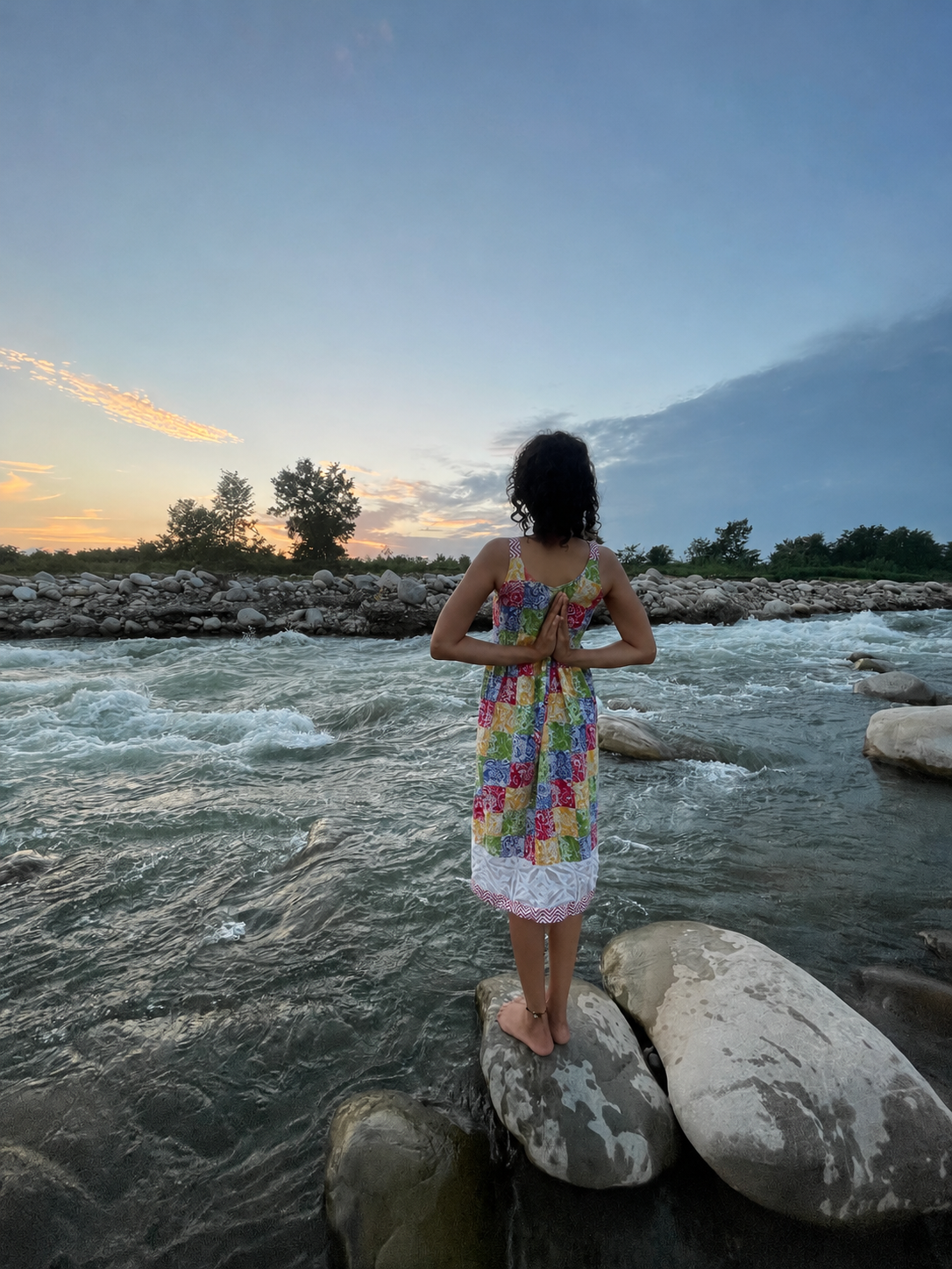 Likhitha Sree in prayer pose at riverside sunset, wearing a patchwork dress