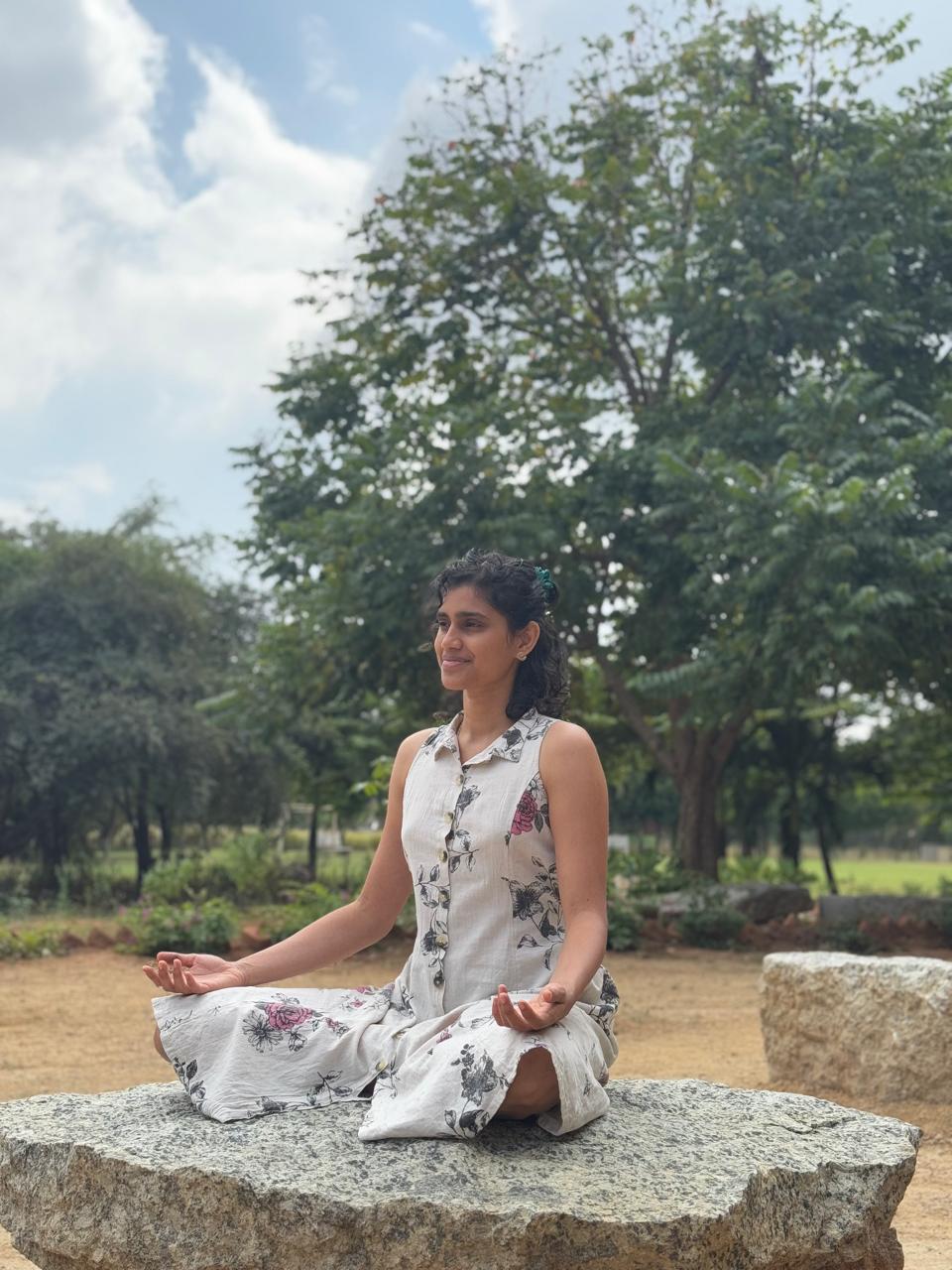 Likhitha in lotus meditation on a flat rock outdoors, serene tree backdrop